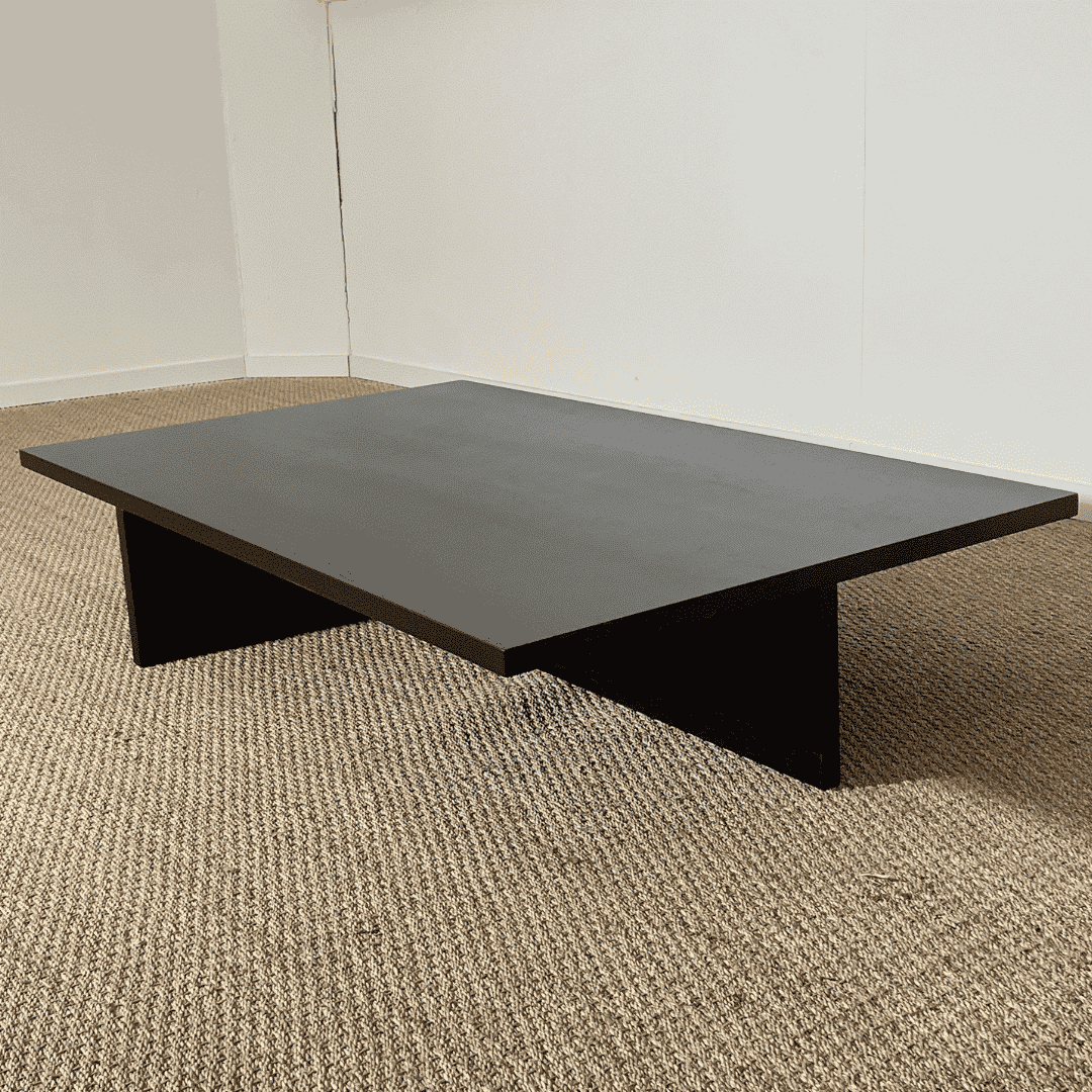 Table basse rectangulaire en placage de chêne teinté noir, avec un effet bois brûlé. Son design minimaliste présente un plateau épais reposant sur une base géométrique formée de panneaux croisés, créant un espace ouvert en dessous. L’ensemble est posé sur un sol en jonc de mer, dans un décor sobre et contemporain.