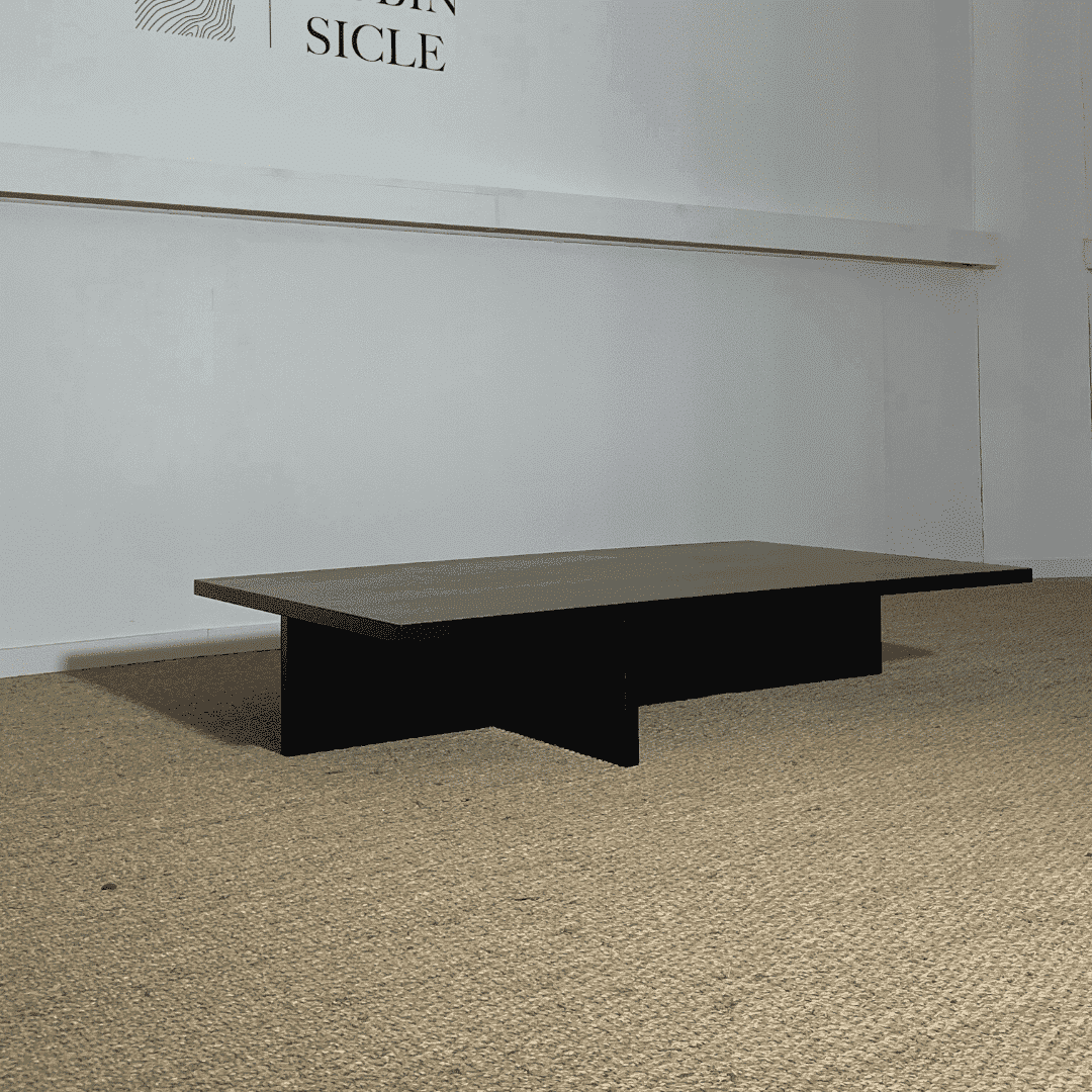 Table basse rectangulaire en placage de chêne teinté noir, avec un effet bois brûlé. Son design minimaliste présente un plateau épais reposant sur une base géométrique formée de panneaux croisés, créant un espace ouvert en dessous. L’ensemble est posé sur un sol en jonc de mer, dans un décor sobre et contemporain.