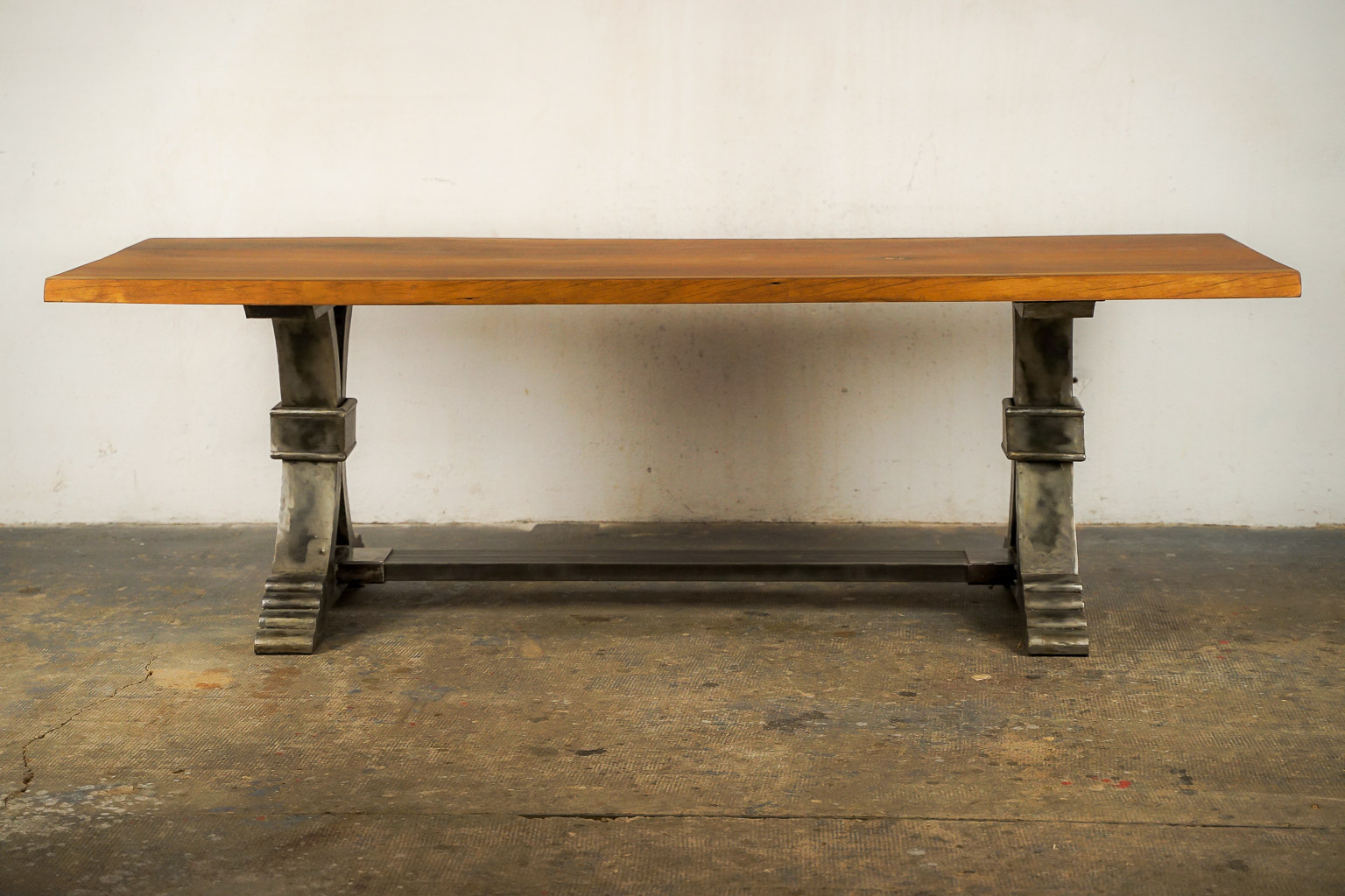 Table à manger au style industriel et élégant, dotée d’un plateau en bois massif teinté chaud. Le piétement central en métal brossé adopte une forme architecturale avec des courbes croisées et une base évasée à marches, évoquant la robustesse des structures anciennes. Le contraste entre le bois chaleureux et le métal brut confère à l’ensemble un caractère à la fois raffiné et authentique.