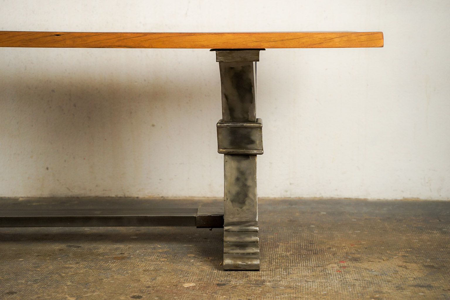 Table à manger au style industriel et élégant, dotée d’un plateau en bois massif teinté chaud. Le piétement central en métal brossé adopte une forme architecturale avec des courbes croisées et une base évasée à marches, évoquant la robustesse des structures anciennes. Le contraste entre le bois chaleureux et le métal brut confère à l’ensemble un caractère à la fois raffiné et authentique.