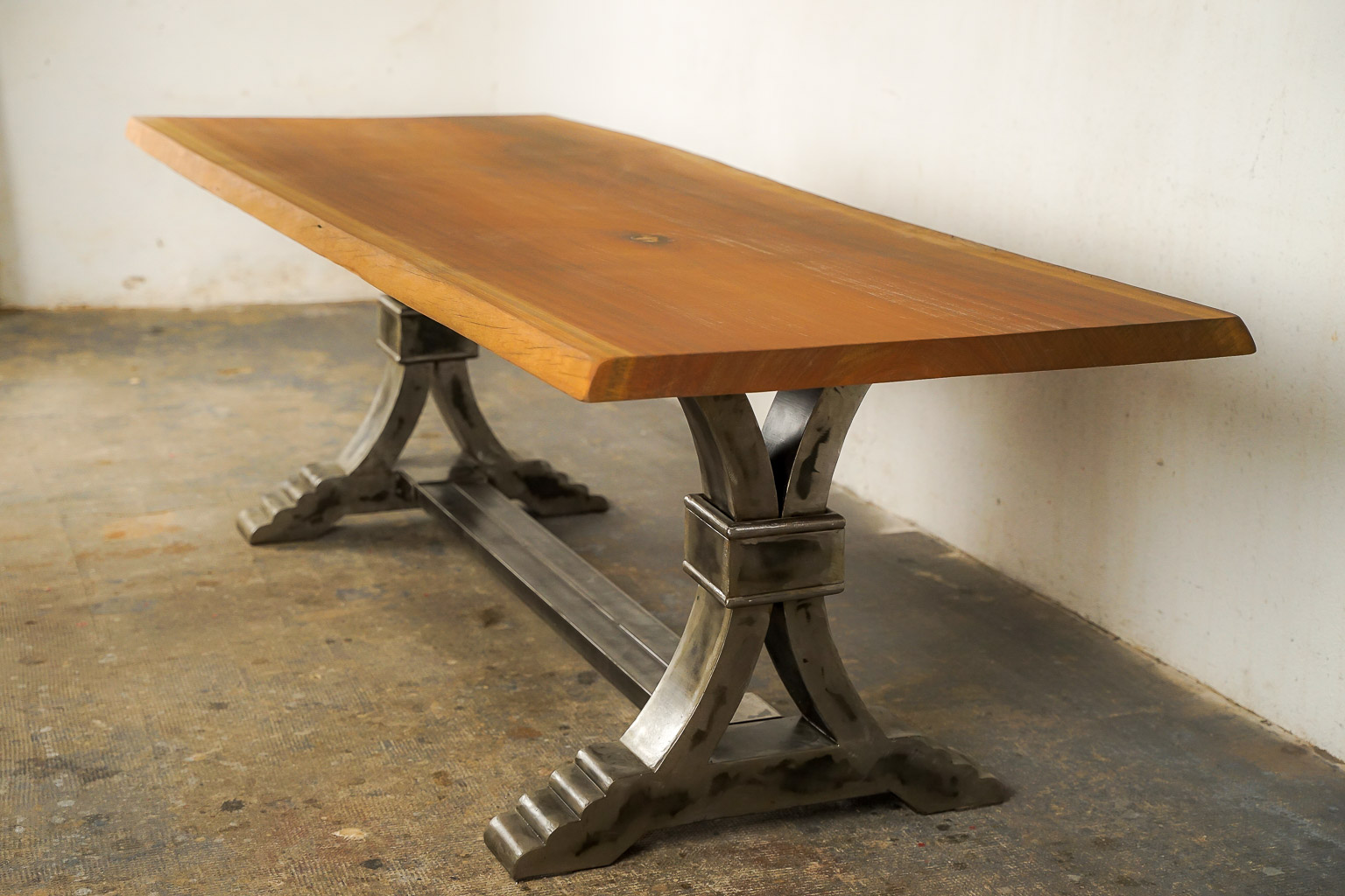 Table à manger au style industriel et élégant, dotée d’un plateau en bois massif teinté chaud. Le piétement central en métal brossé adopte une forme architecturale avec des courbes croisées et une base évasée à marches, évoquant la robustesse des structures anciennes. Le contraste entre le bois chaleureux et le métal brut confère à l’ensemble un caractère à la fois raffiné et authentique.