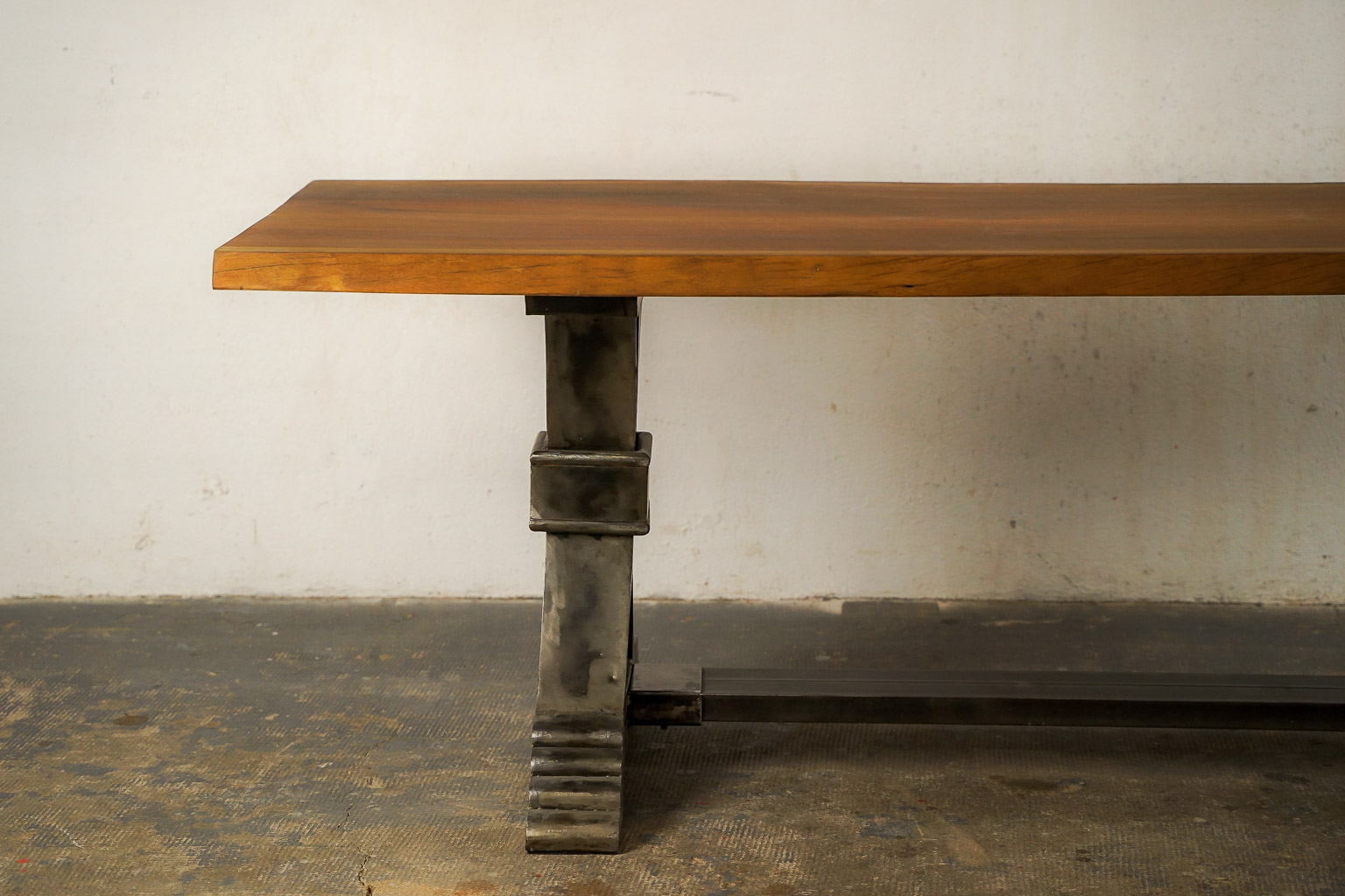 Table à manger au style industriel et élégant, dotée d’un plateau en bois massif teinté chaud. Le piétement central en métal brossé adopte une forme architecturale avec des courbes croisées et une base évasée à marches, évoquant la robustesse des structures anciennes. Le contraste entre le bois chaleureux et le métal brut confère à l’ensemble un caractère à la fois raffiné et authentique.