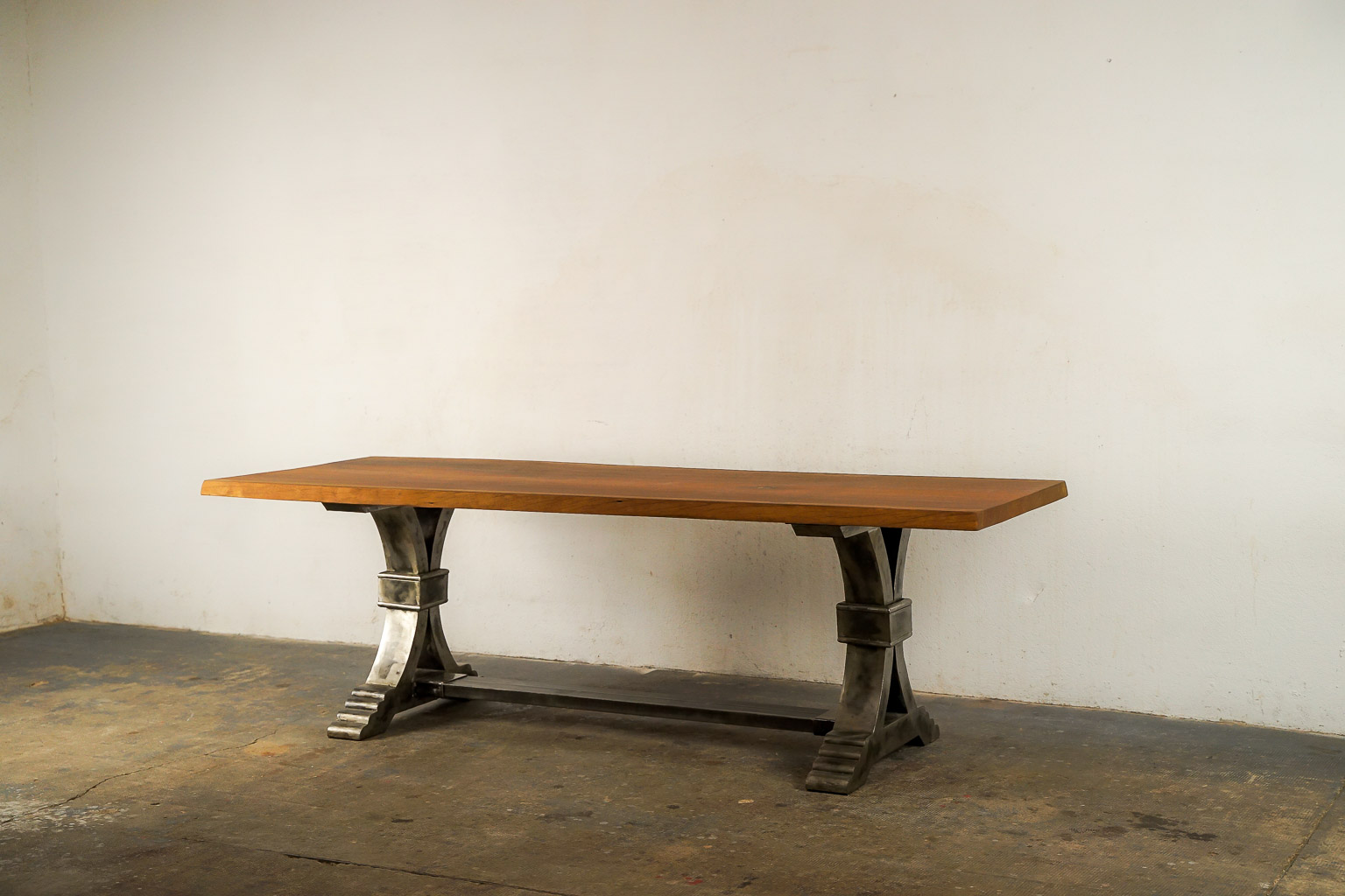 Table à manger au style industriel et élégant, dotée d’un plateau en bois massif teinté chaud. Le piétement central en métal brossé adopte une forme architecturale avec des courbes croisées et une base évasée à marches, évoquant la robustesse des structures anciennes. Le contraste entre le bois chaleureux et le métal brut confère à l’ensemble un caractère à la fois raffiné et authentique.