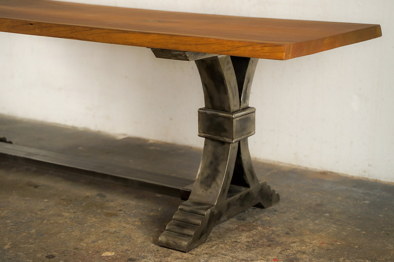 Table à manger au style industriel et élégant, dotée d’un plateau en bois massif teinté chaud. Le piétement central en métal brossé adopte une forme architecturale avec des courbes croisées et une base évasée à marches, évoquant la robustesse des structures anciennes. Le contraste entre le bois chaleureux et le métal brut confère à l’ensemble un caractère à la fois raffiné et authentique.