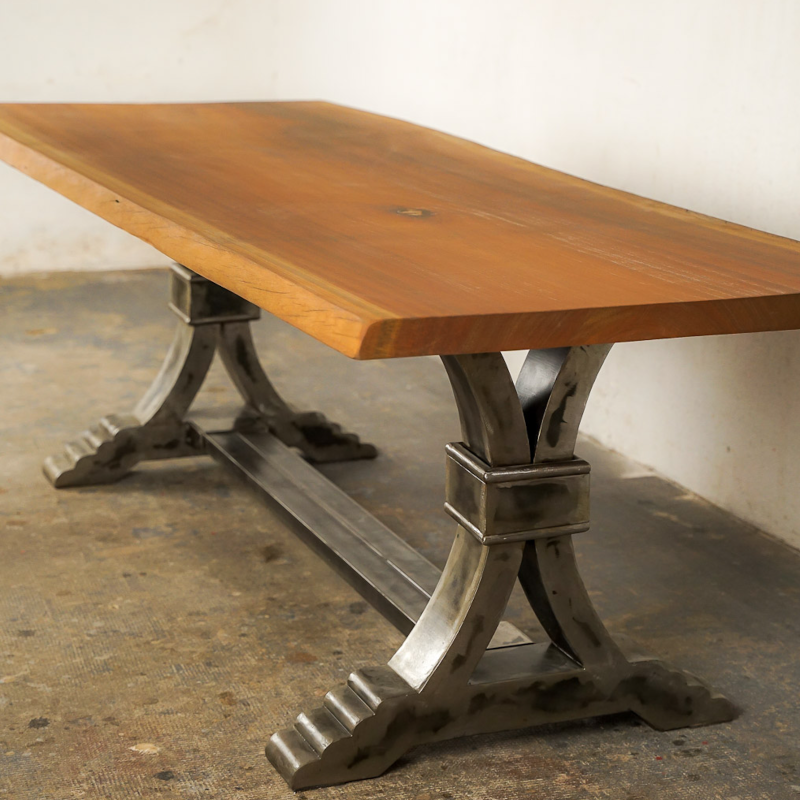 Table à manger au style industriel et élégant, dotée d’un plateau en bois massif teinté chaud. Le piétement central en métal brossé adopte une forme architecturale avec des courbes croisées et une base évasée à marches, évoquant la robustesse des structures anciennes. Le contraste entre le bois chaleureux et le métal brut confère à l’ensemble un caractère à la fois raffiné et authentique.